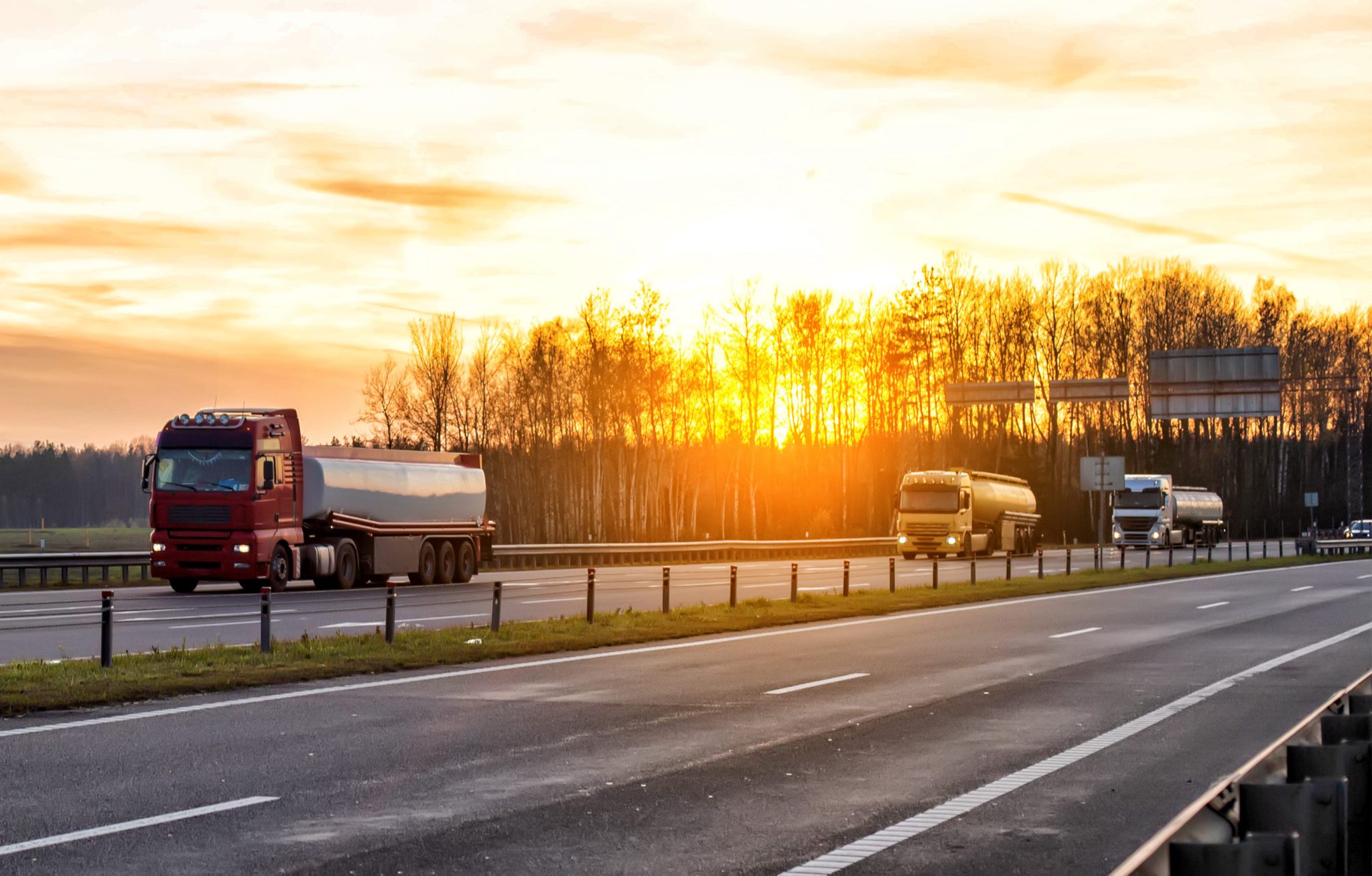 A convoy of three trucks transports dangerous goods in tank semi-trailers on a motorway against the backdrop of sunset in the evening.